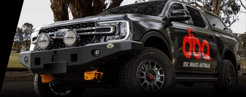 Pickup truck oscuro con branding DBA Disc Brakes Australia con logo DBA rojo y equipo iluminación adicional en entorno exterior