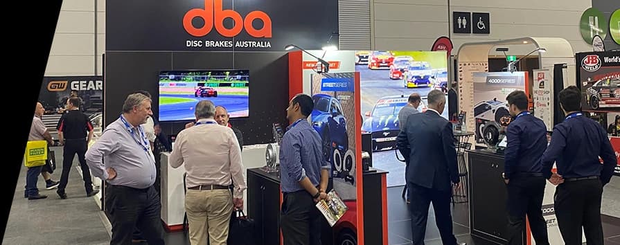 Stand de exposición DBA en feria automotriz con logotipo de la empresa destacado y asistentes viendo exhibiciones de productos y contenido de motorsport