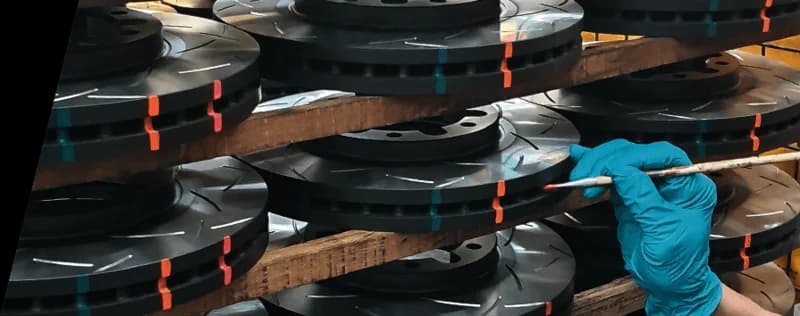 Técnico con guantes azules inspeccionando discos de freno ranurados DBA apilados con marcas indicadoras de temperatura durante control de calidad