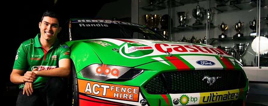 Piloto de carreras sonriente con camisa verde de equipo junto a Ford Mustang verde patrocinado por DBA con múltiples logotipos de patrocinadores en sala de trofeos