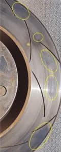 Disco de freno con depósitos visibles de material de fricción que pueden provocar vibración del volante y frenado irregular Primer plano de la superficie de un disco de freno mostrando depósitos irregulares de material de fricción resaltados con círculos amarillos