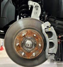 Paso 1A: Conjunto de freno original mostrando disco y pinza que serán reemplazados durante el procedimiento de actualización DBA Primer plano del conjunto de freno original mostrando superficie de disco oxidada, pinza y soporte de montaje antes de la instalación de la actualización DBA
